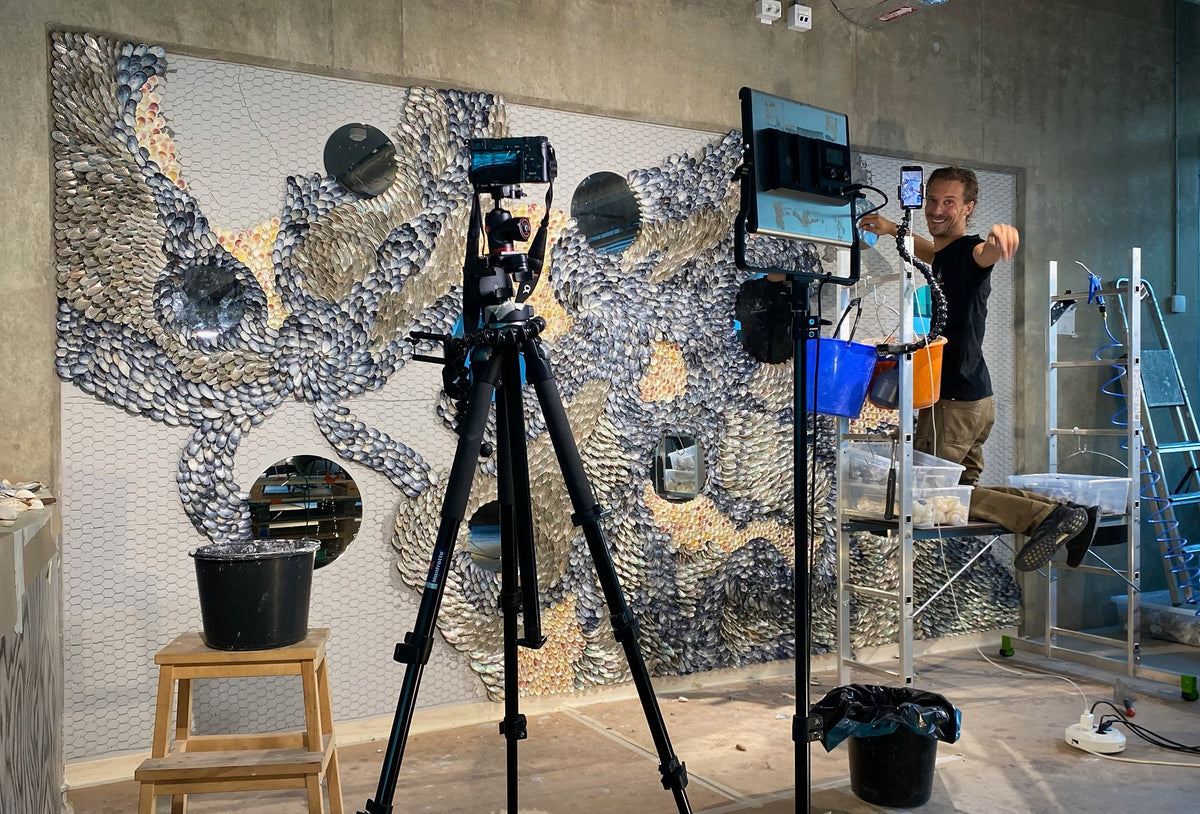 Cameras and a light on tripods filming the half-finished shell wall for a time-lapse, Buster Eriksson smiling on the scaffold